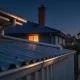 A suburban Australian roofline at dusk, with a corrugated metal roof in the foreground and tiled roofs and lit windows along the street.