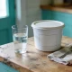 A glass of water beside a plain unlabelled white tub on a timber kitchen bench in soft morning light.