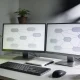 Two monitors showing heavily blurred flow diagrams on a home office desk with a keyboard, a notebook and a coffee.