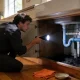 A person crouching at an open sink cabinet shining a torch on the pipework beneath the basin in an Australian kitchen.