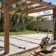 A half-built timber pergola frame over a concrete slab with a ladder, a mitre saw and timber offcuts in an Australian backyard.
