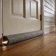 A worn grey draught excluder against the base of a pale timber front door in an older Australian hallway, with daylight showing where the seal fails.