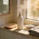 A pale tiled ledge in a warm Australian bathroom with an unbranded pump bottle, a tanning mitt, a folded towel and a dish of moisturiser in afternoon light.