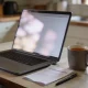 An open laptop angled away on a kitchen bench, its screen a soft blurred glow, beside a mug of tea, a pen and a folded statement.