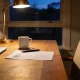 A quiet home office corner in the early evening with a lit lamp over a table holding a printed page, pen, mug and a closed laptop.