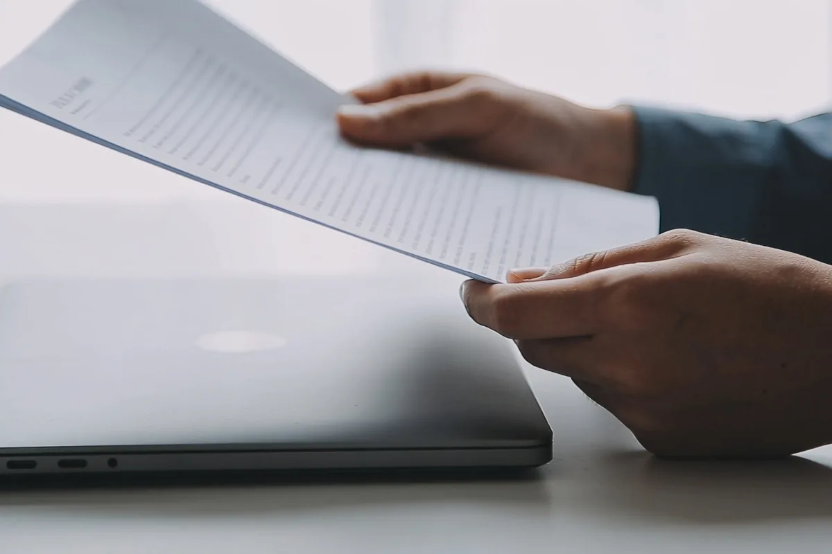Two hands holding a printed document above a desk, about to set it down beside a closed silver laptop.