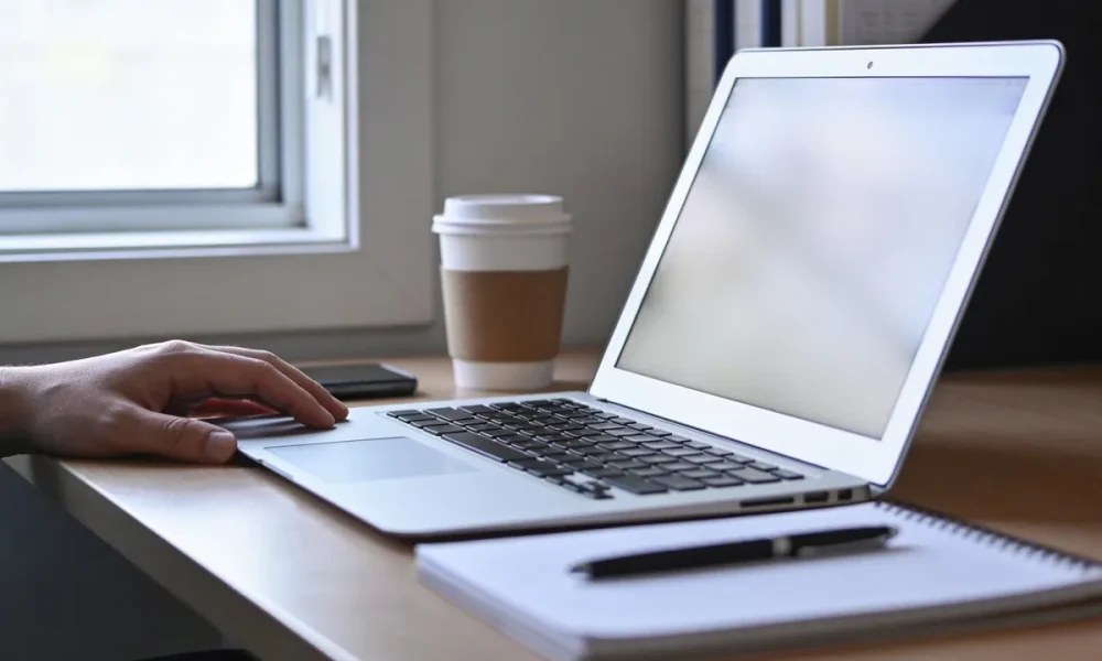 A desk in mid-morning light with an open laptop showing a blank screen, a notepad, a coffee cup, a phone face down and a hand resting beside the laptop.