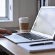 A desk in mid-morning light with an open laptop showing a blank screen, a notepad, a coffee cup, a phone face down and a hand resting beside the laptop.