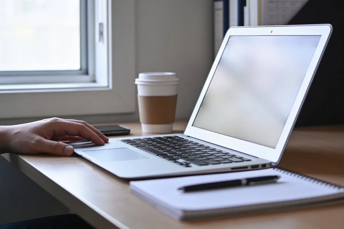 A desk in mid-morning light with an open laptop showing a blank screen, a notepad, a coffee cup, a phone face down and a hand resting beside the laptop.