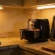An air fryer with its drawer open and steam rising on a kitchen bench beside a baking tray, tea towel and chopping board in warm overhead light.