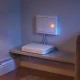A plain white wall-mounted connection box with a glowing amber light above a white modem on a low shelf, cables clipped along the skirting in an evening living room.