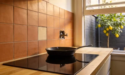 A black induction cooktop set into a timber benchtop with a cast-iron frying pan on it, a capped old gas fitting and one mismatched tile in the terracotta splashback, and a lemon tree in the courtyard outside