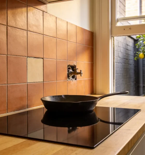 A black induction cooktop set into a timber benchtop with a cast-iron frying pan on it, a capped old gas fitting and one mismatched tile in the terracotta splashback, and a lemon tree in the courtyard outside