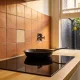 A black induction cooktop set into a timber benchtop with a cast-iron frying pan on it, a capped old gas fitting and one mismatched tile in the terracotta splashback, and a lemon tree in the courtyard outside
