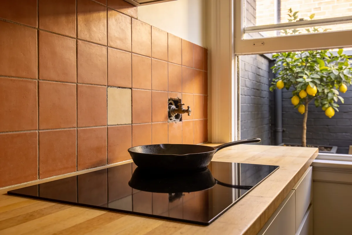 A black induction cooktop set into a timber benchtop with a cast-iron frying pan on it, a capped old gas fitting and one mismatched tile in the terracotta splashback, and a lemon tree in the courtyard outside