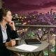 A woman in work clothes at a small table on an apartment balcony at dusk with a mug, a closed laptop and a sheet of paper face down, a flowering jacaranda and city lights below