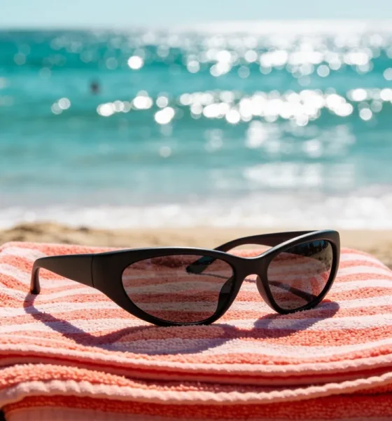 Black wraparound sunglasses resting on a folded coral and white striped towel on the sand, with sun glare sparkling on the sea behind