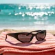 Black wraparound sunglasses resting on a folded coral and white striped towel on the sand, with sun glare sparkling on the sea behind