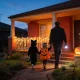 Two small children dressed as a black cat and a pumpkin walking hand in hand with an adult carrying a flashlight up the path to a brick house with a glowing carved pumpkin and fairy lights at dusk