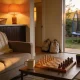 A wooden chess set and two mugs on a coffee table in a family lounge at dusk, with a knitted throw on the couch, two school bags by the hallway and a backyard trampoline through the open sliding door