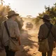 Two young fruit pickers in wide-brimmed hats and long sleeves walking between rows of orange trees with canvas picking bags, a farm ute raising dust behind them in early morning light