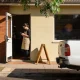 A cafe owner in a canvas apron checks her phone in the open doorway of a small brick shopfront on a sunny suburban street, with a gum tree and a white ute at the kerb.