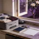 A half-packed carry-on bag on a kitchen bench beside a closed navy travel document, a phone and a blank sheet of paper, with a frangipani tree outside the open window in late-afternoon light.