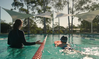 A swim instructor stands waist-deep in an outdoor pool guiding a young child in a swim cap kicking with a kickboard, under shade sails with eucalypts beyond the fence.