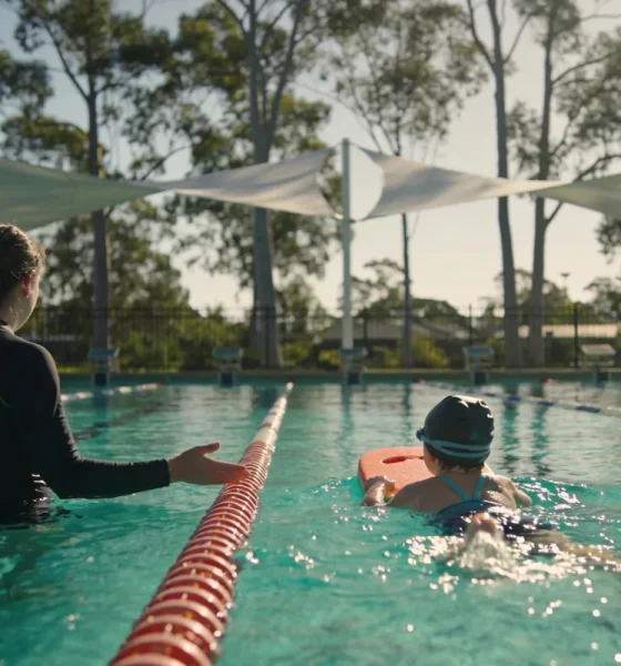 A swim instructor stands waist-deep in an outdoor pool guiding a young child in a swim cap kicking with a kickboard, under shade sails with eucalypts beyond the fence.
