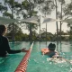 A swim instructor stands waist-deep in an outdoor pool guiding a young child in a swim cap kicking with a kickboard, under shade sails with eucalypts beyond the fence.