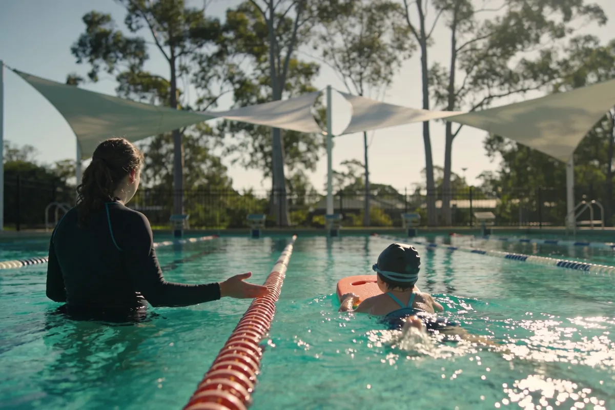 A swim instructor stands waist-deep in an outdoor pool guiding a young child in a swim cap kicking with a kickboard, under shade sails with eucalypts beyond the fence.